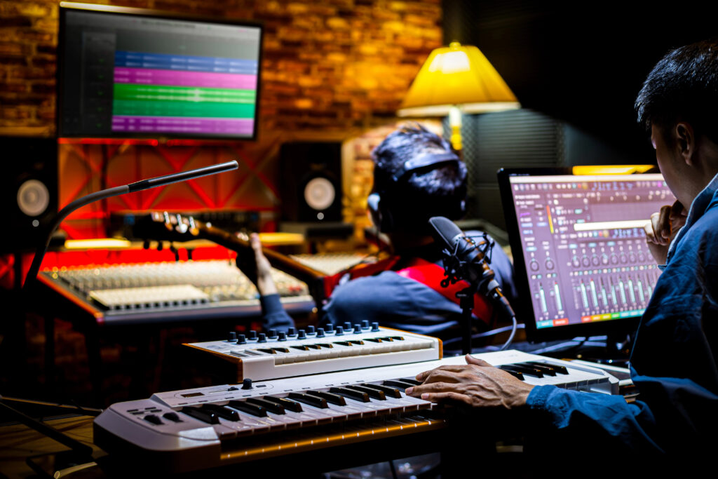 Producteurs travaillant en studio d’enregistrement avec clavier MIDI, guitare et logiciels MAO, illustrant le travail de ghost producer et la formation DJ en ligne.